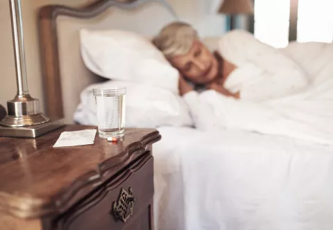 Un verre d'eau et des médicaments, avec en arrière plan, une femme âgée qui dort